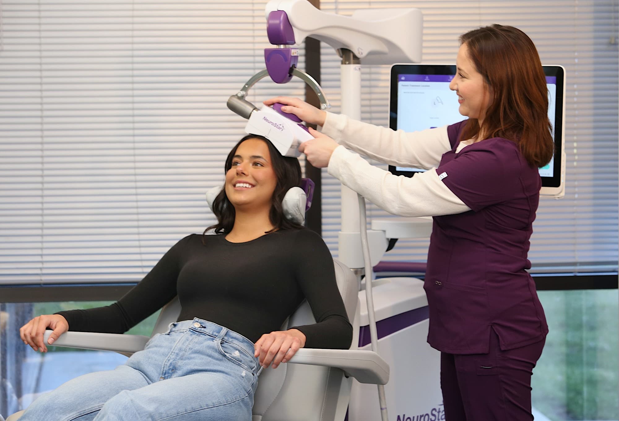 A smiling patient sits back in a NeuroStar TMS treatment chair while a care team member positions the treatment coil